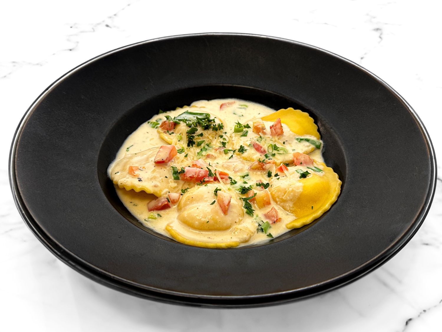 Ravioli in a creamy white sauce with diced tomatoes and herbs, served in a wide-rimmed black bowl on a white surface.