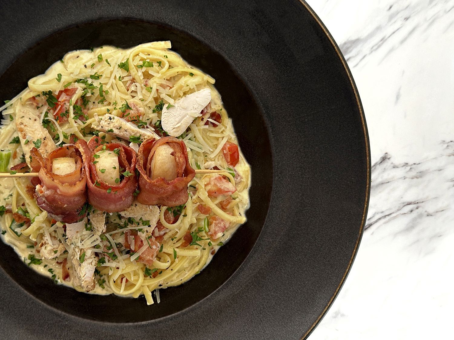 A bowl of creamy pasta topped with three bacon-wrapped scallops on a skewer, served on a black plate with a marble table.