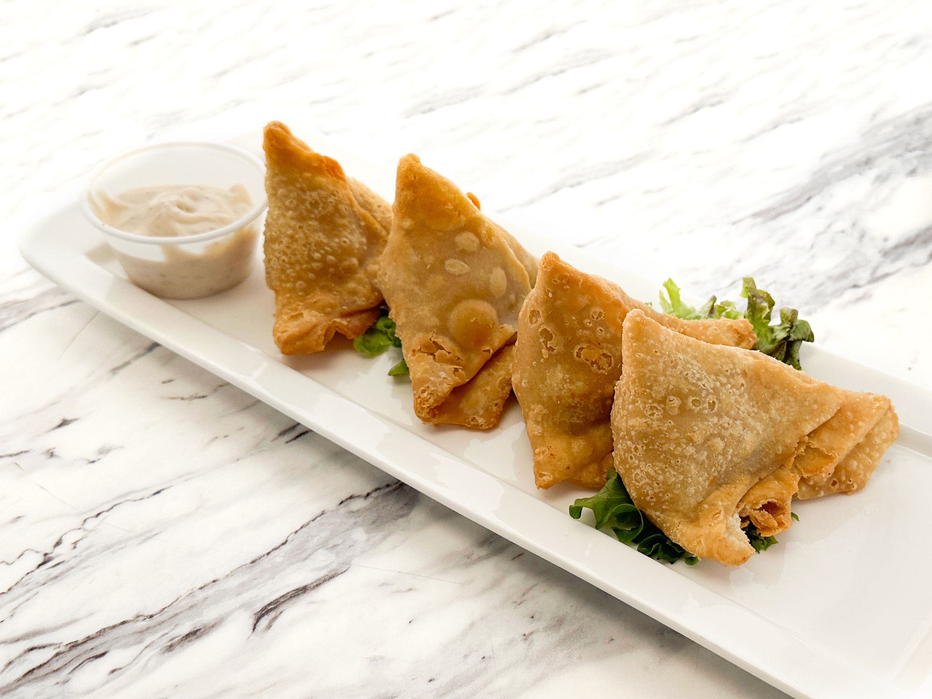 Four golden-brown fried samosas arranged on a white rectangular platter with a side of dipping sauce.