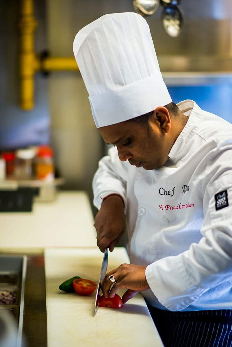 A chef in a white uniform and tall hat focuses on slicing a red pepper on a white cutting board in a professional kitchen.