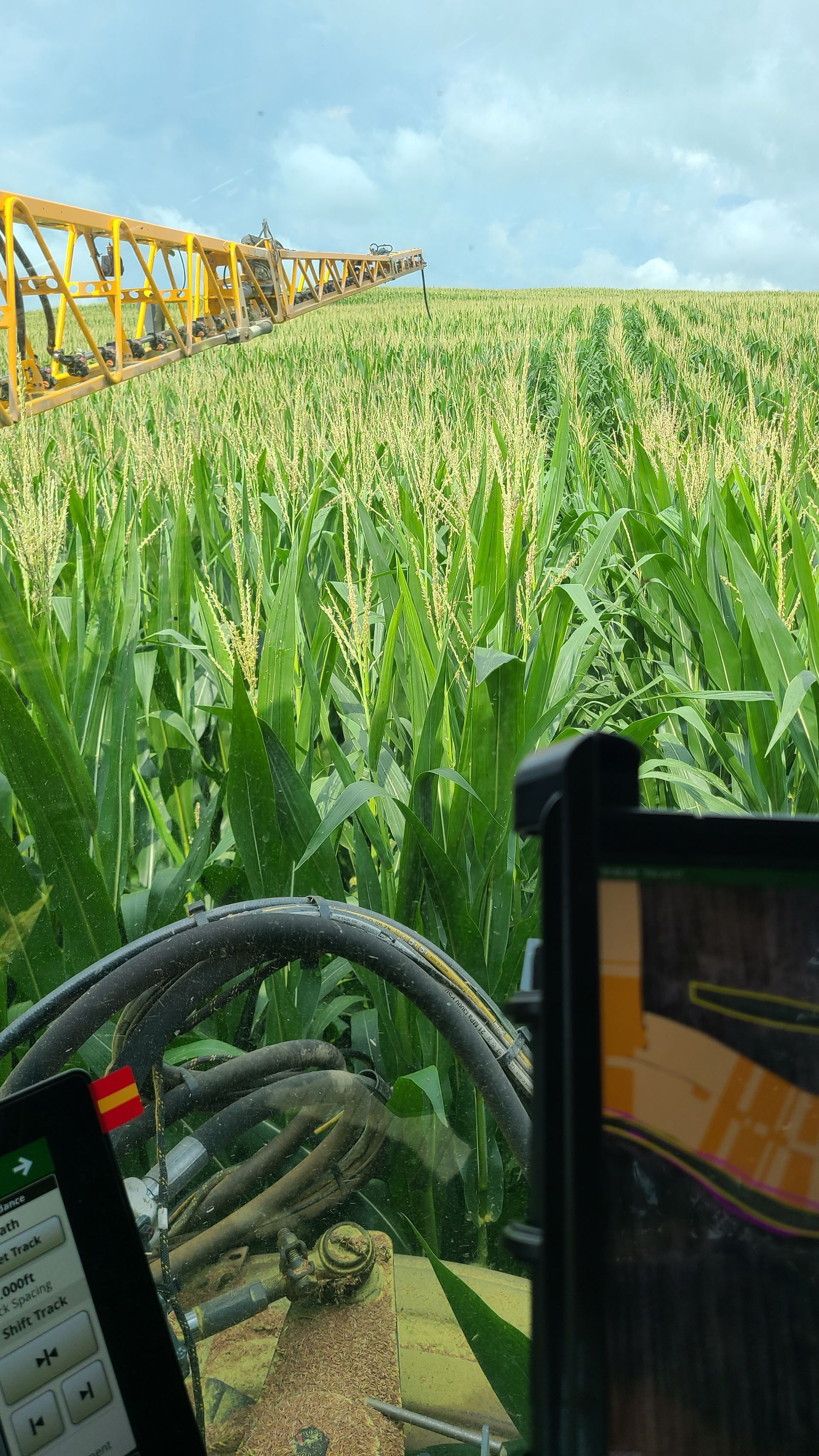 View from a tractor cab showing the long yellow boom of a sprayer extending over a dense green cornfield under a cloudy sky.