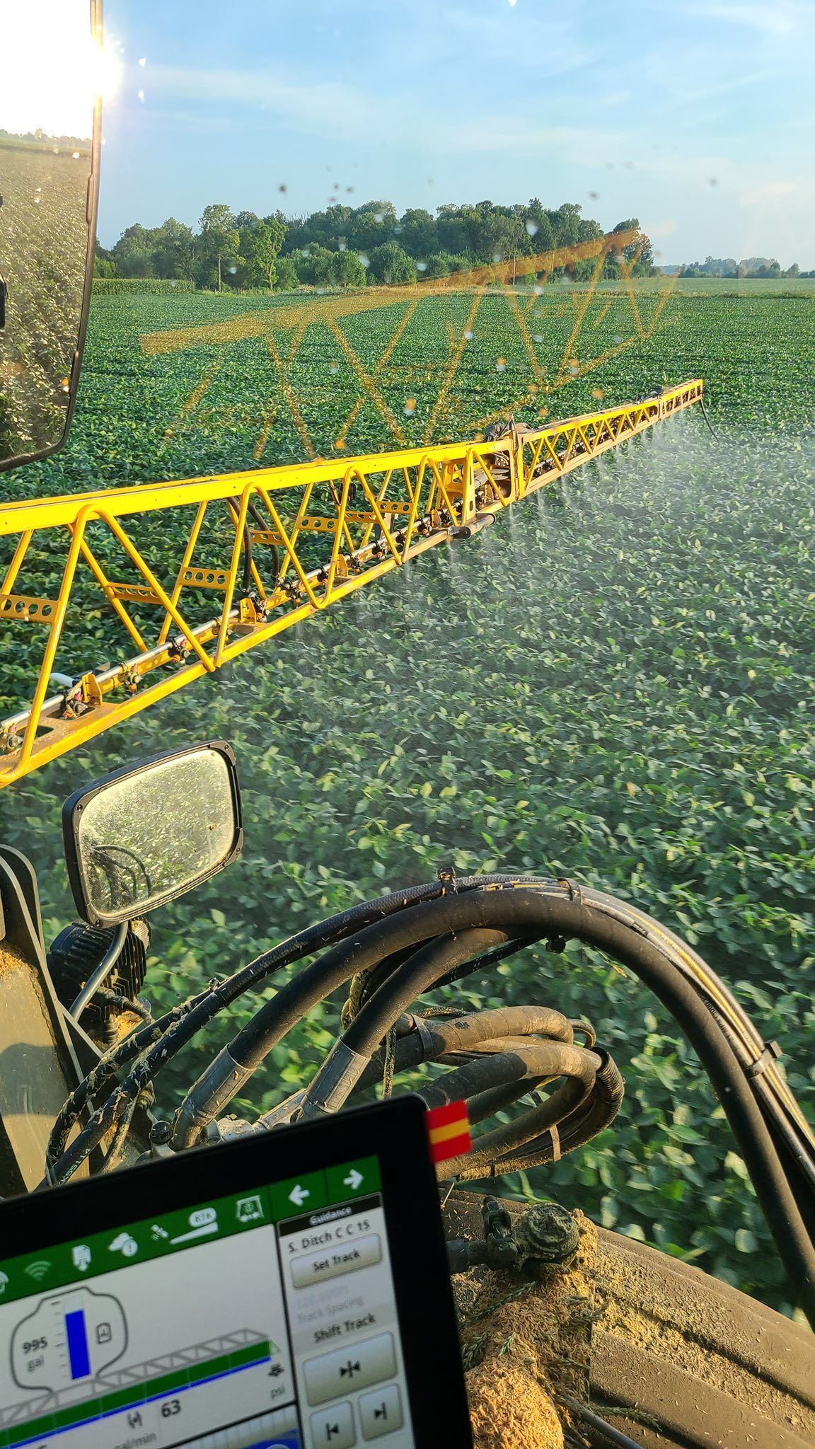 View from a tractor cab showing a yellow sprayer boom applying mist to a large field of green crops under a sunny sky.