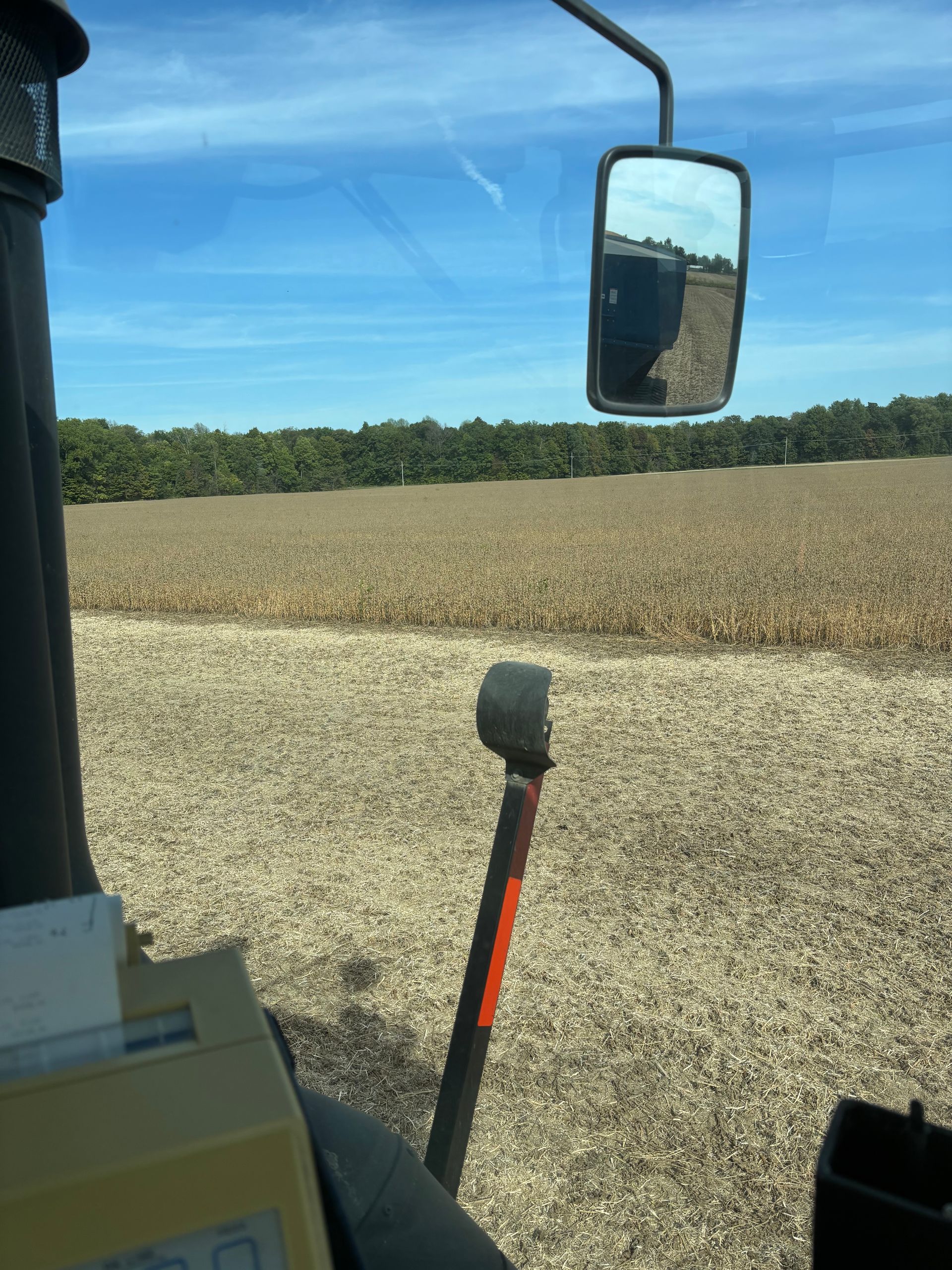 View from inside a tractor cab looking out at a harvested field with trees in the distance under a clear blue sky.