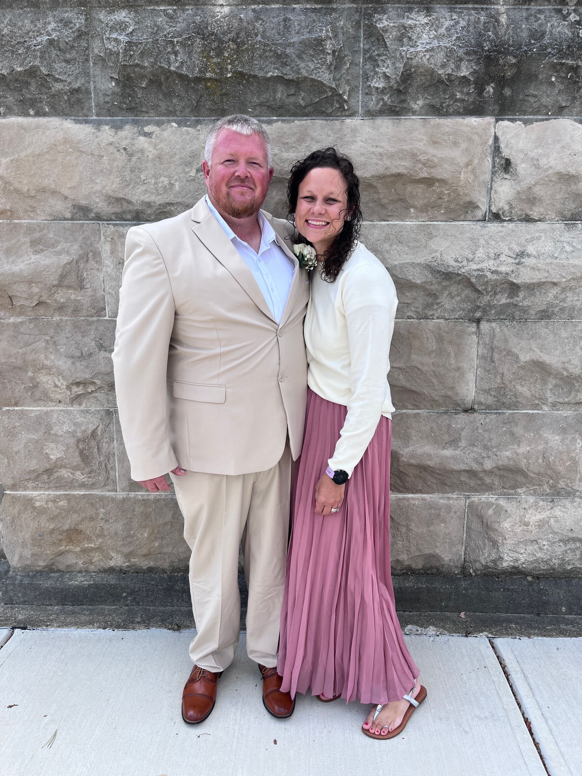 A couple poses for a photo in front of a stone wall; the man wears a beige suit, and the woman wears a cream top and pink skirt.