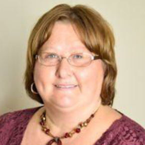 Smiling woman in glasses wearing a purple lace top and beaded necklace against a plain background