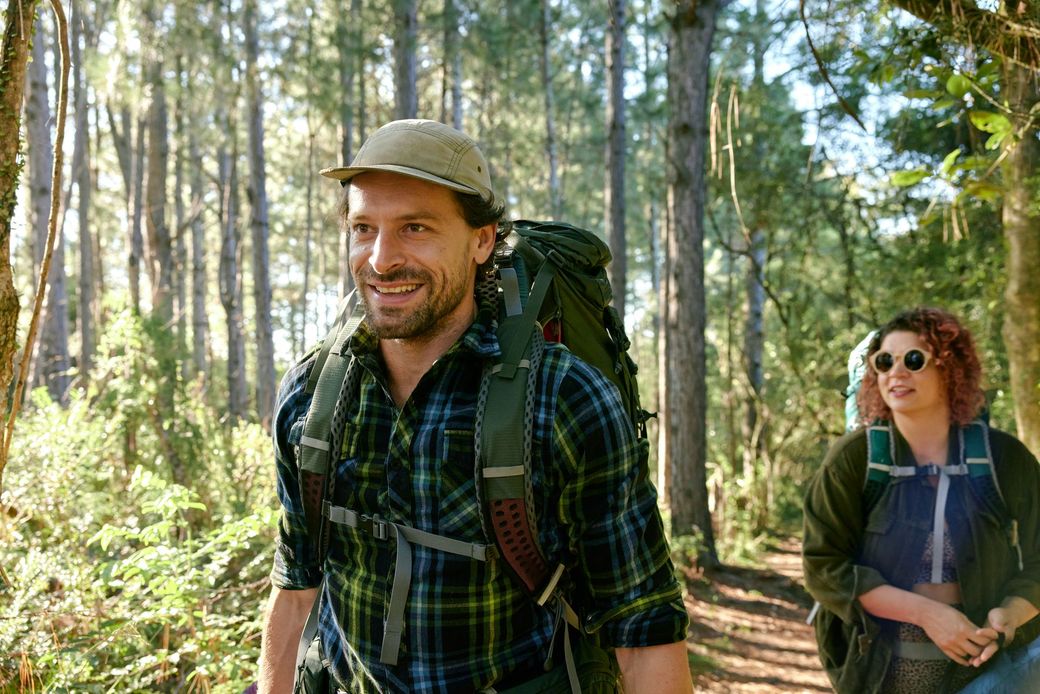Two hikers with backpacks walking through a sunlit forest, smiling and wearing outdoor gear.