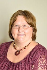 Portrait of a smiling person wearing glasses, a maroon lace top, and a beaded necklace against a plain background