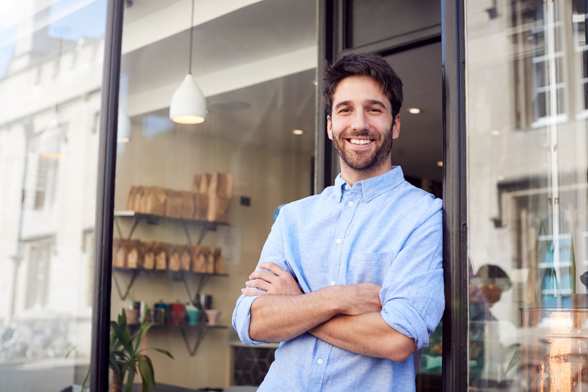 Smiling man in a light blue shirt stands with arms crossed outside a shop window