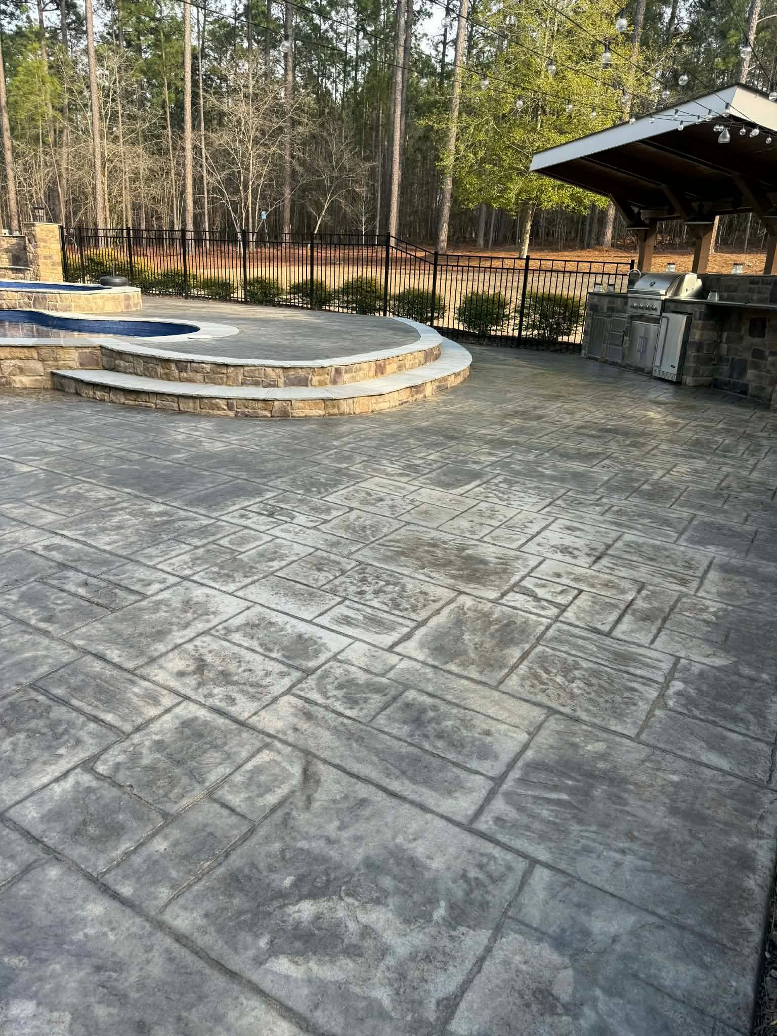 Concrete walkway curves towards a pool. Workers in orange shirts are visible in background. Poolside concrete path under construction, leading to a blue pool, surrounded by dirt. Two workers in the background.