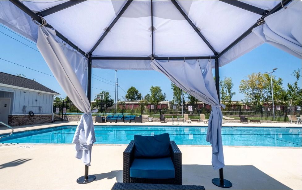 view of the swimming pool from inside a cabana with white drapes, cloth roof and an empty blue armchair