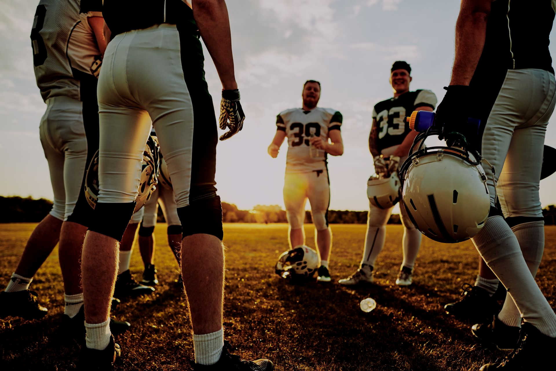 A group of American football players in uniforms standing in a huddle on a field during a golden sunset.