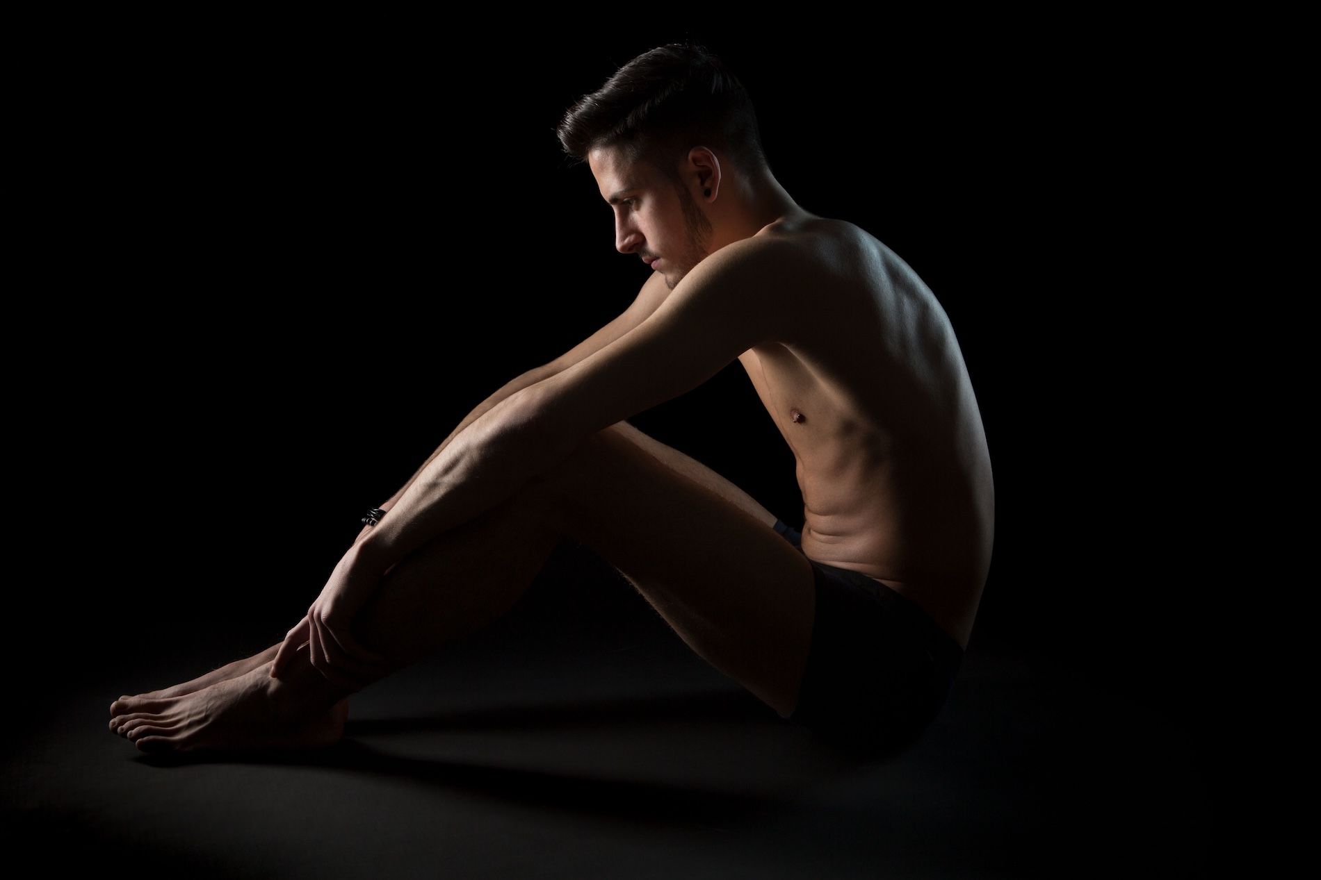 A person sits on a dark background in profile, with their head bowed and hands resting on their knees.