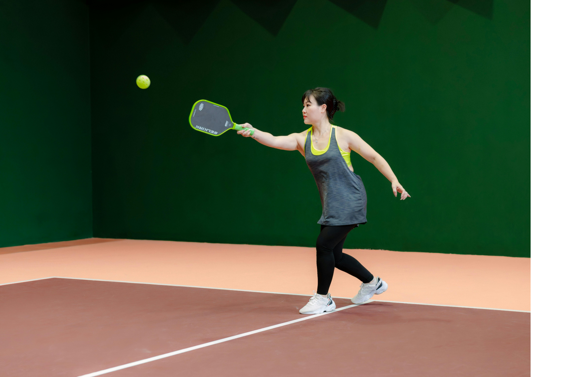 A person in a gray tank top and black leggings hits a neon yellow pickleball with a paddle on a court with green walls.