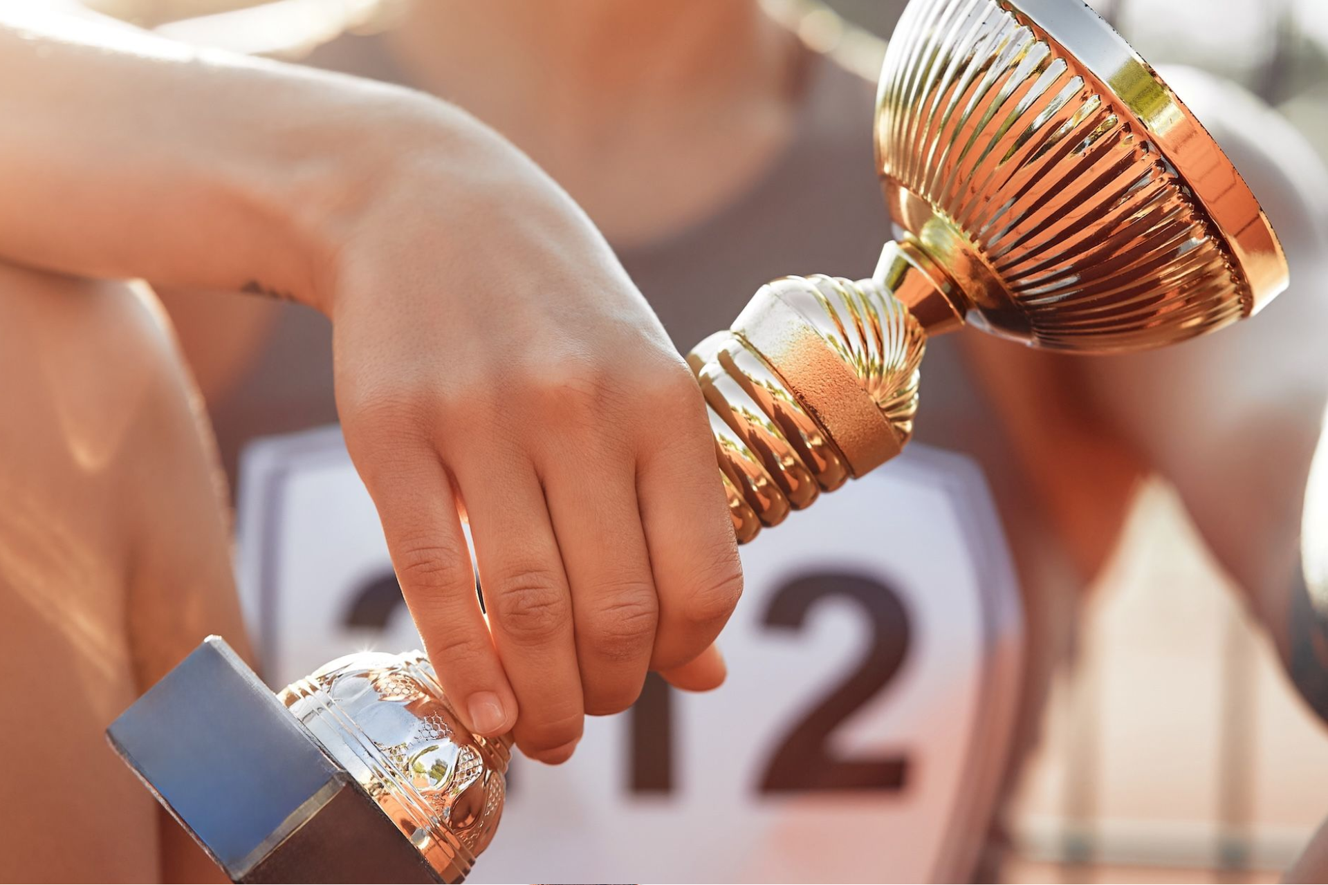 Close-up of a person wearing a bib numbered 12, holding a gold trophy on a running track.