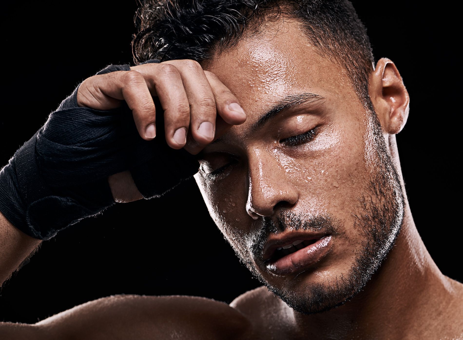 A person with sweat-drenched skin, eyes closed, and wearing athletic hand wraps, pausing against a black background.