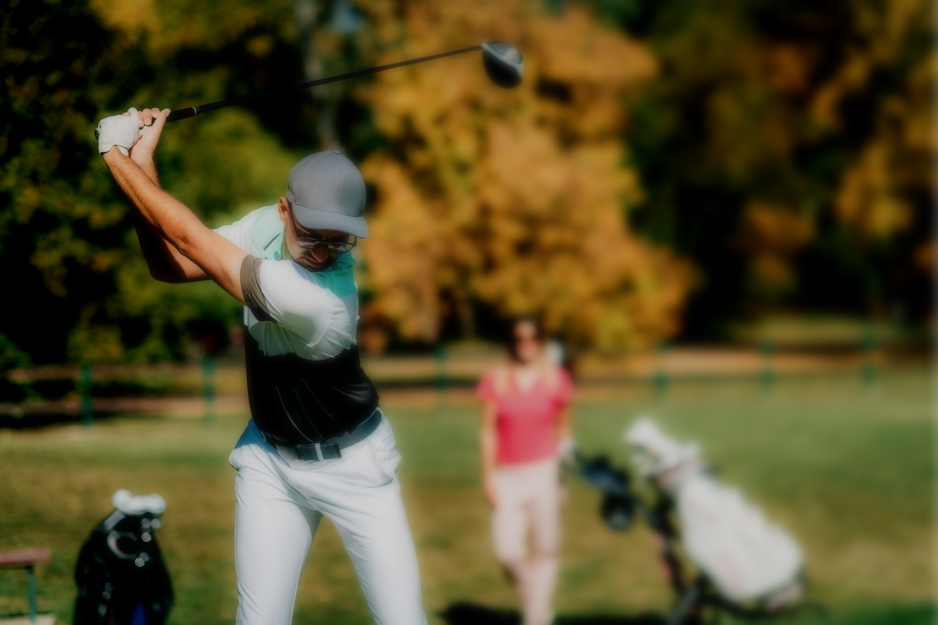 A golfer wearing a cap and light pants at the top of their swing on a sunny, autumn golf course.