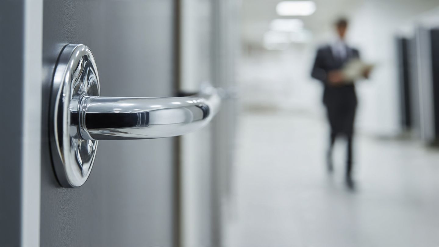 Close-up of a silver door handle with a blurred person standing in a hallway.