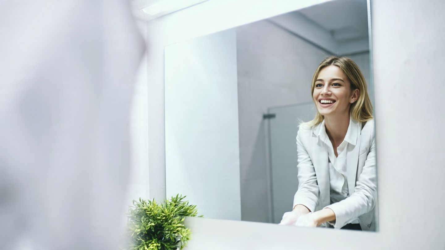Smiling woman in a white blazer leaning on a bathroom counter, seen in a mirror with a small plant nearby