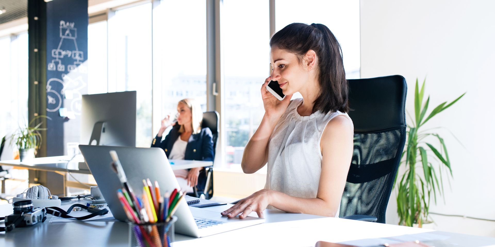 A woman is sitting at a desk in front of a laptop computer talking on a cell phone.