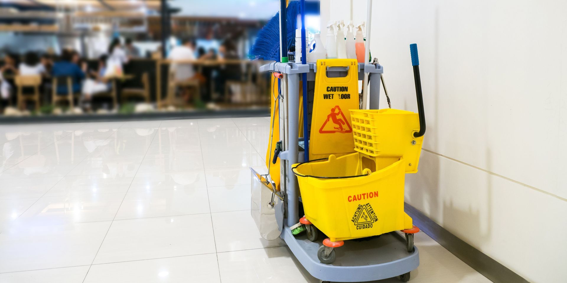 A yellow cleaning cart with a caution sign sits next to a white wall in a public space.