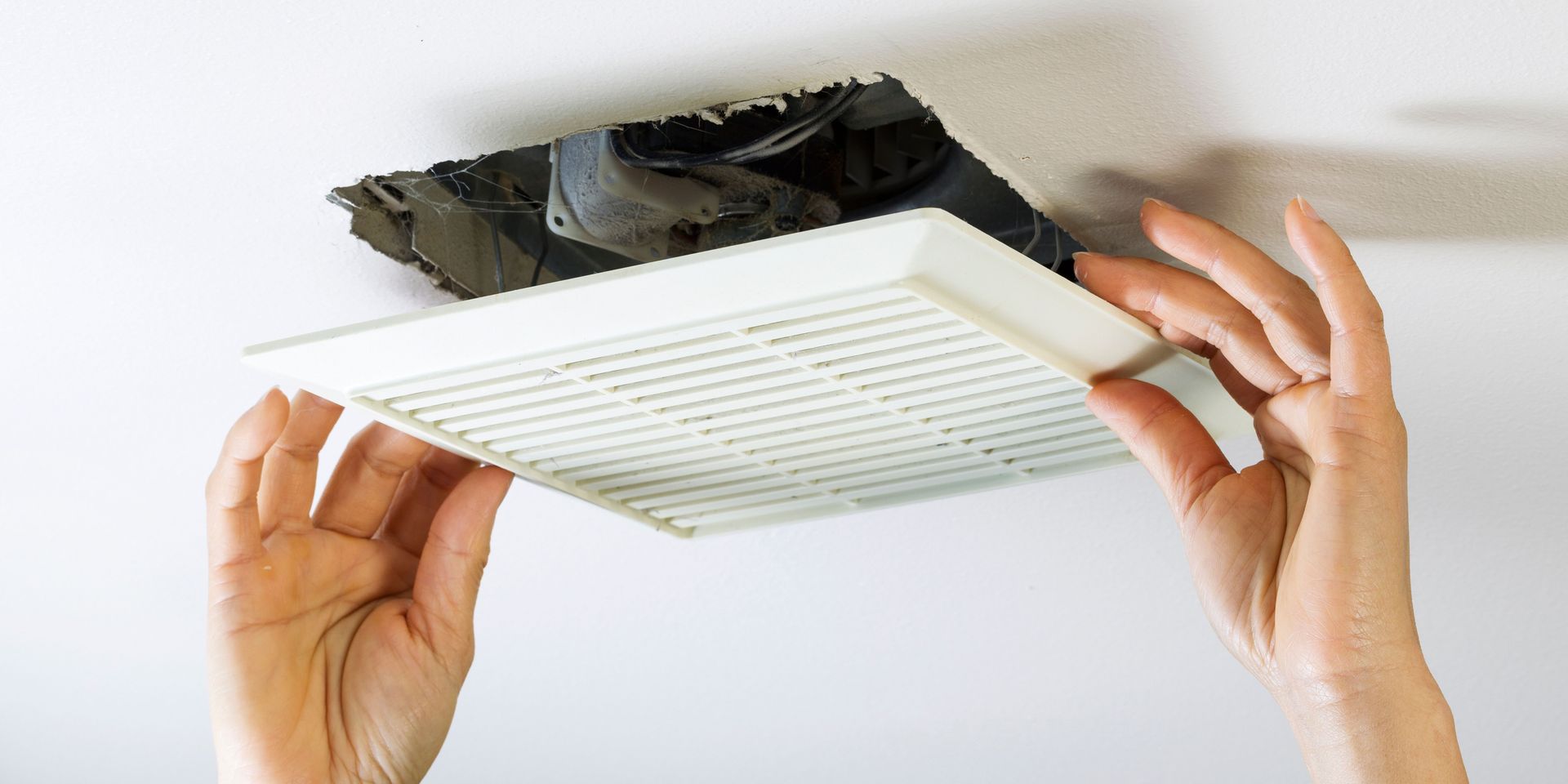 Hands holding a white vent cover up to a hole in a white ceiling.