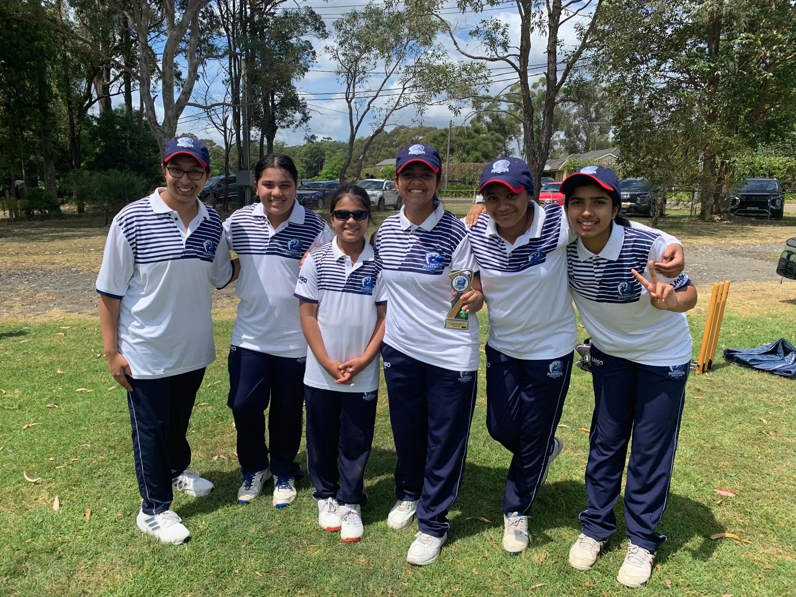 Women's cricket team celebrating a victory on a green field. Players in white uniforms with arms raised, smiling.