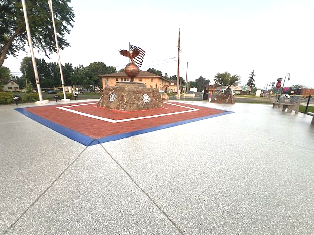 Veterans memorial in Lindstrom, MN in a paved plaza, with a red-white-and-blue border and grey coverings on the outskirt of the concrete