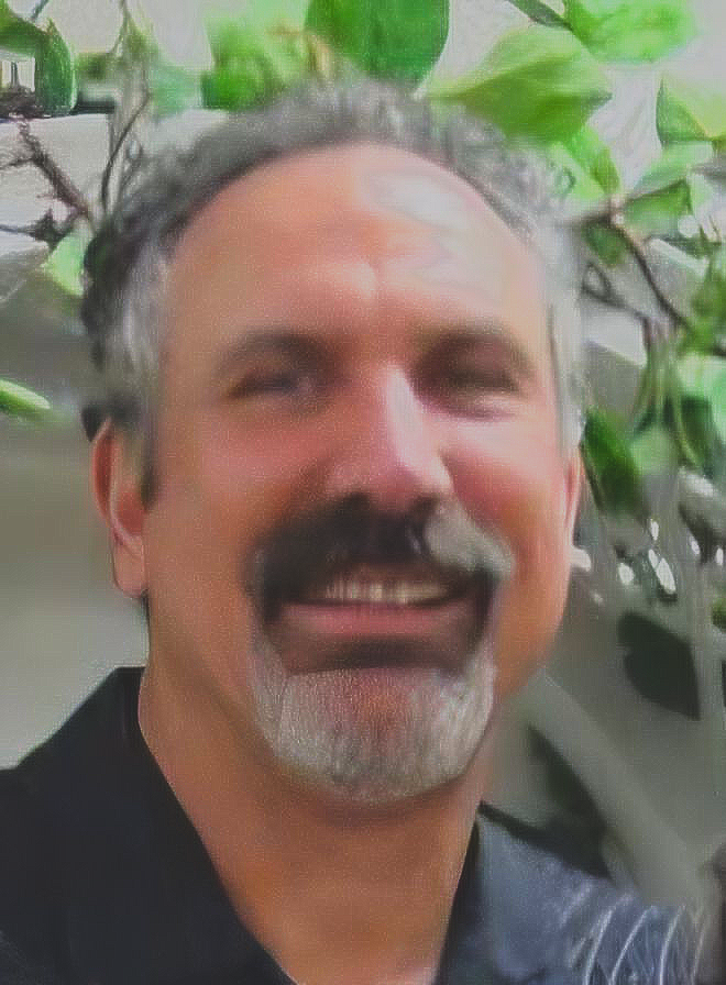 A smiling individual with a dark mustache and graying beard, positioned in front of green foliage.
