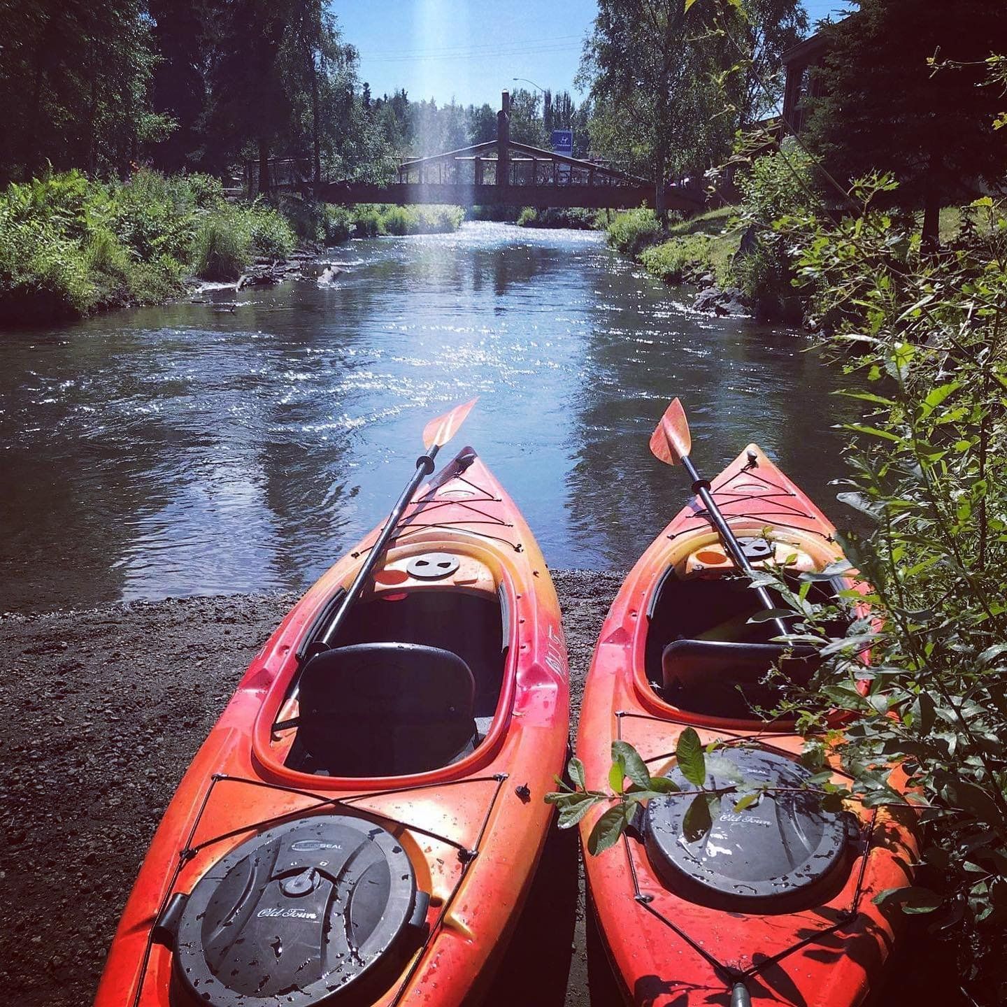 Two orange kayaks on a riverbank, paddles on board, with a bridge and trees in the background.