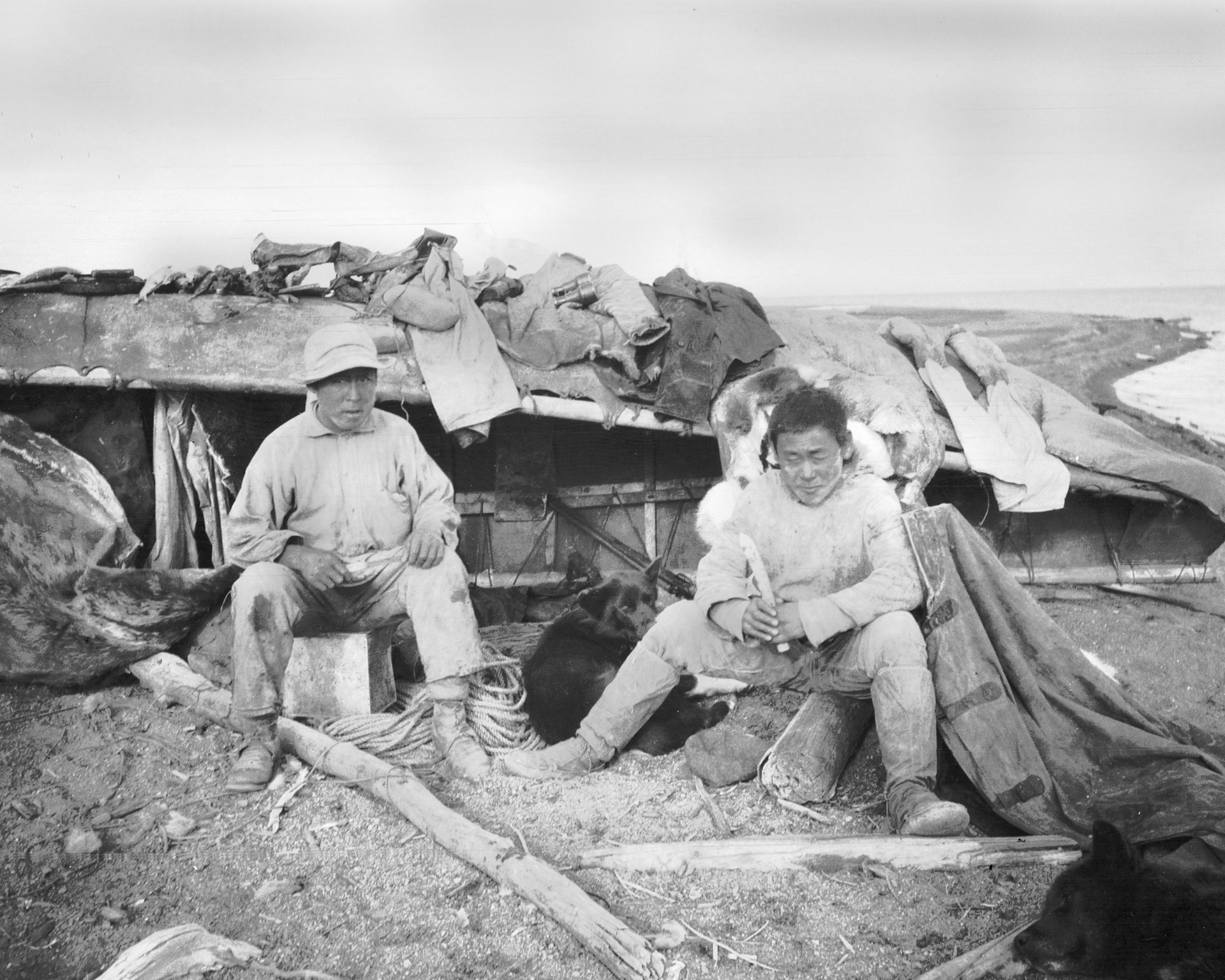 Two people sit outside a low structure. One older person, one child, in front of a coastal scene, black and white photograph.