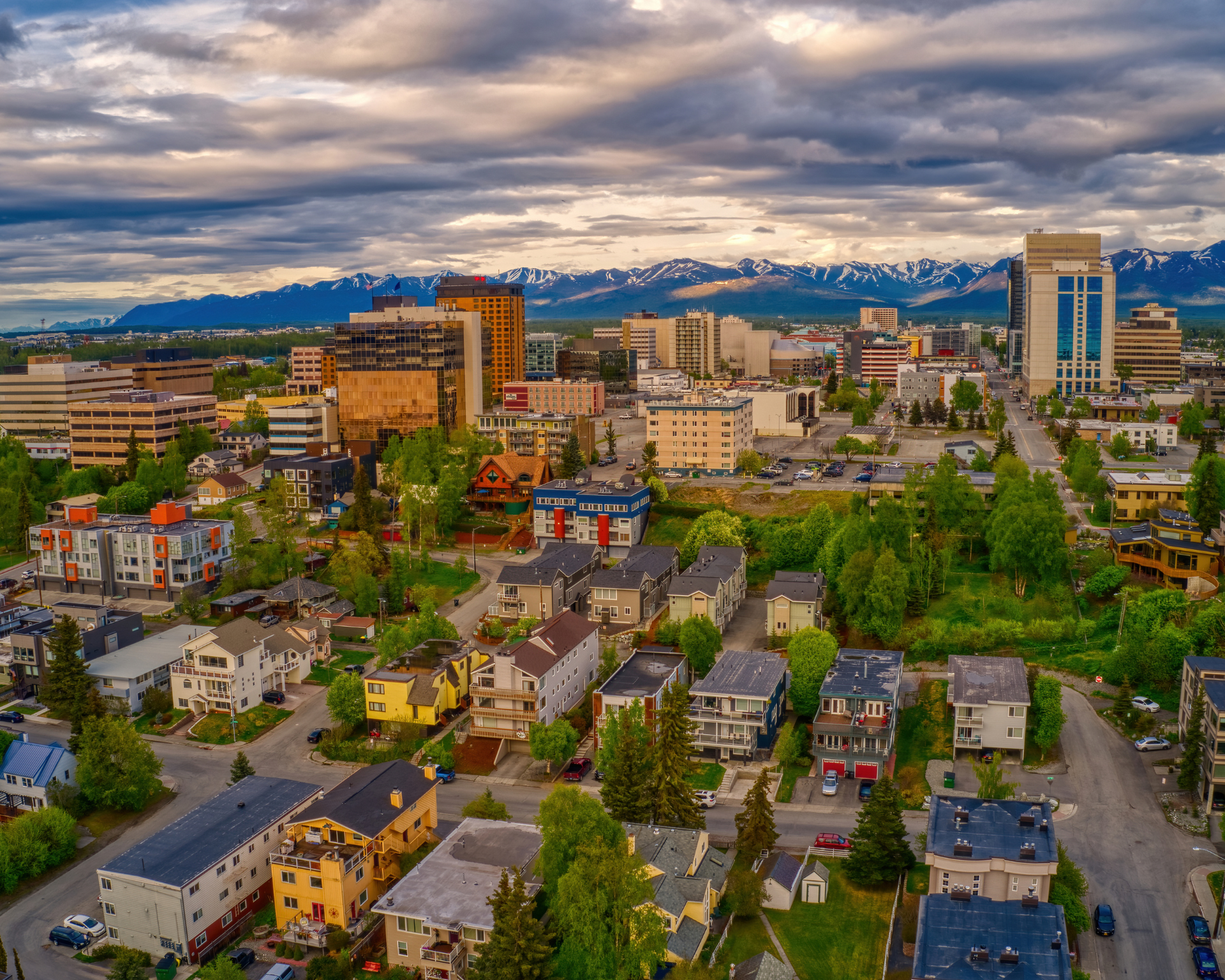 Aerial view of Anchorage, Alaska. Cityscape with buildings, homes, and distant snow-capped mountains under a cloudy sky.