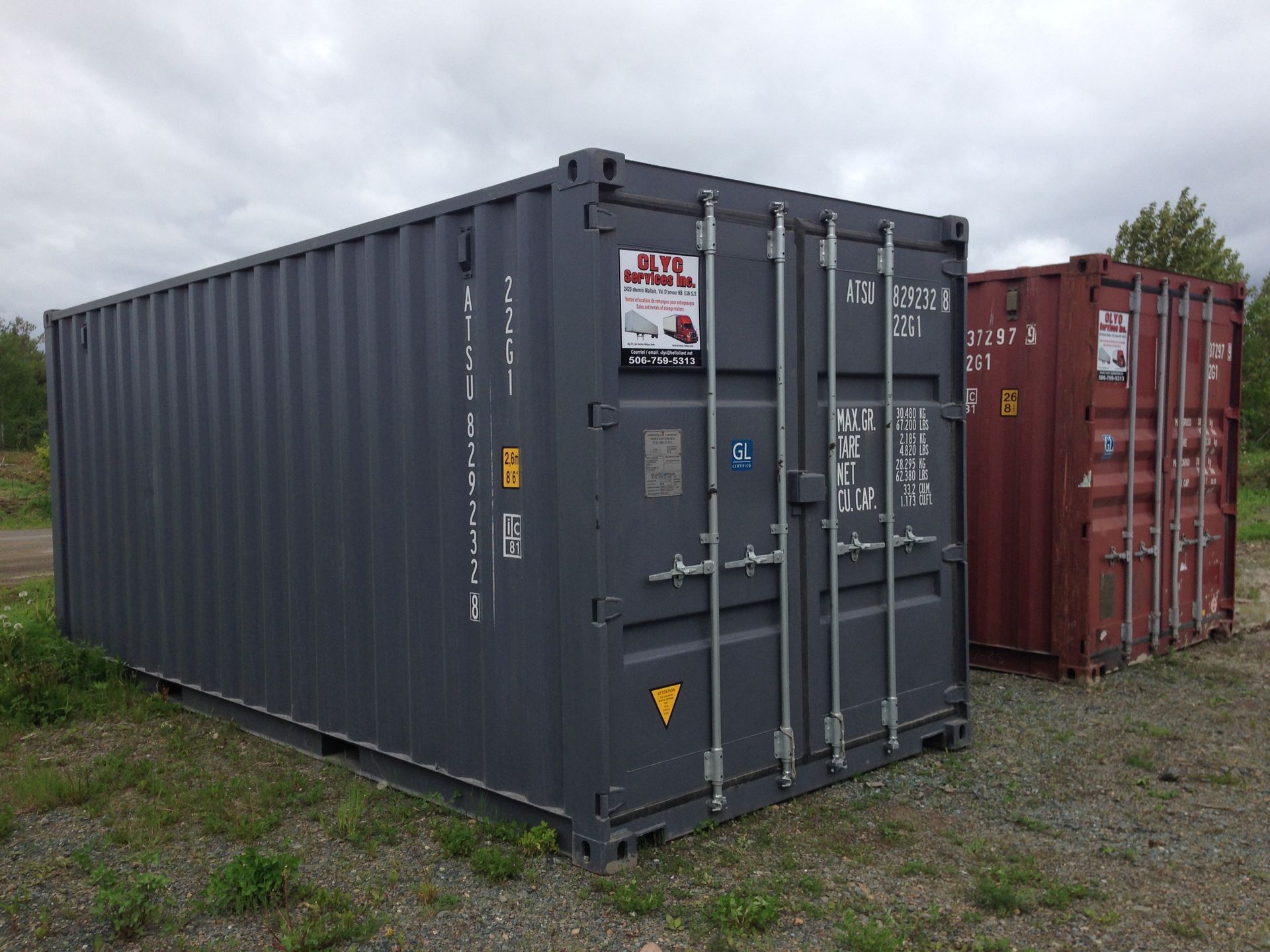 A dark grey shipping container stands on a gravel lot next to a smaller, maroon shipping container under a cloudy sky.