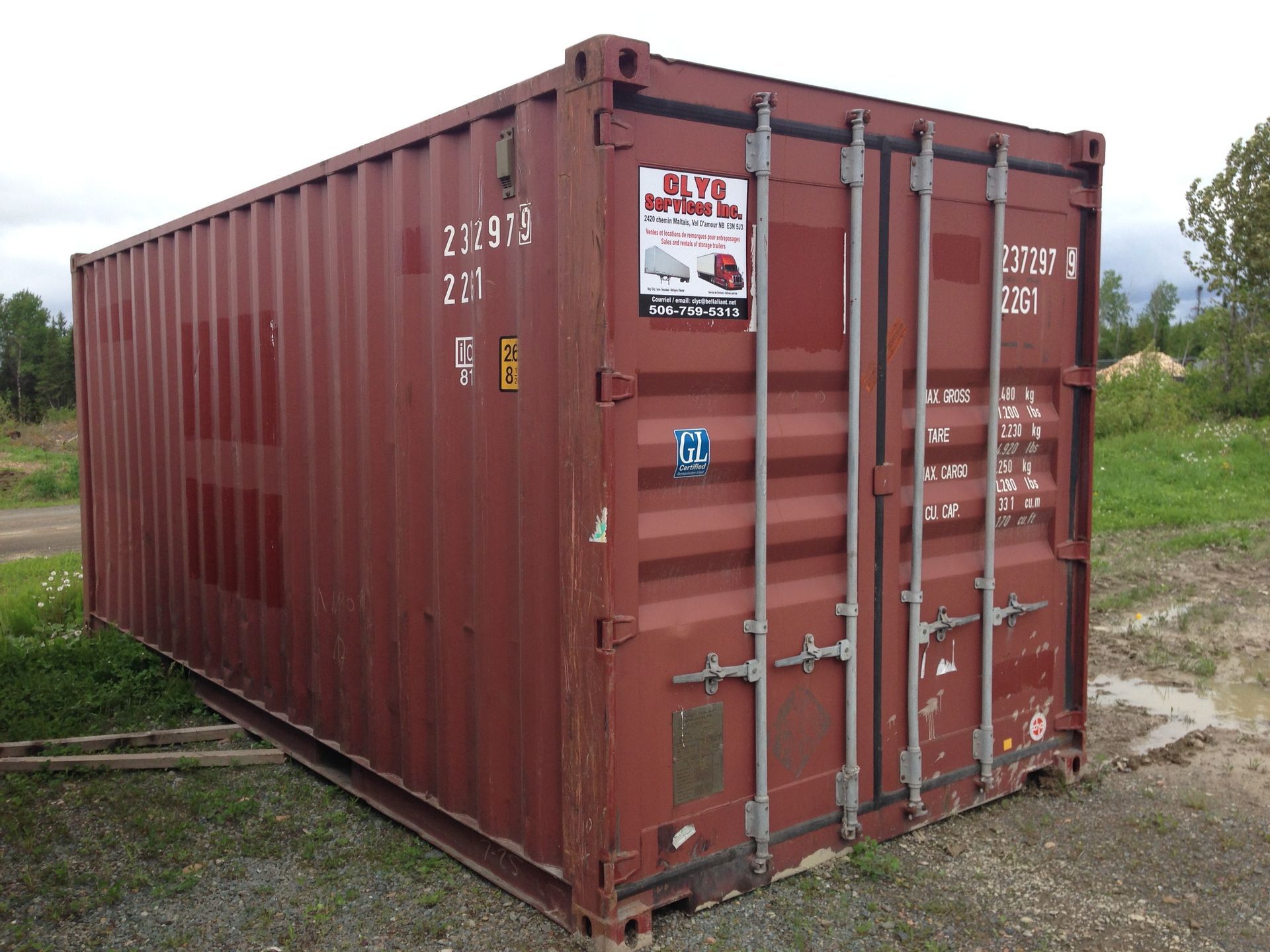 A red shipping container stands on a gravel surface outdoors with a sticker on its closed door.