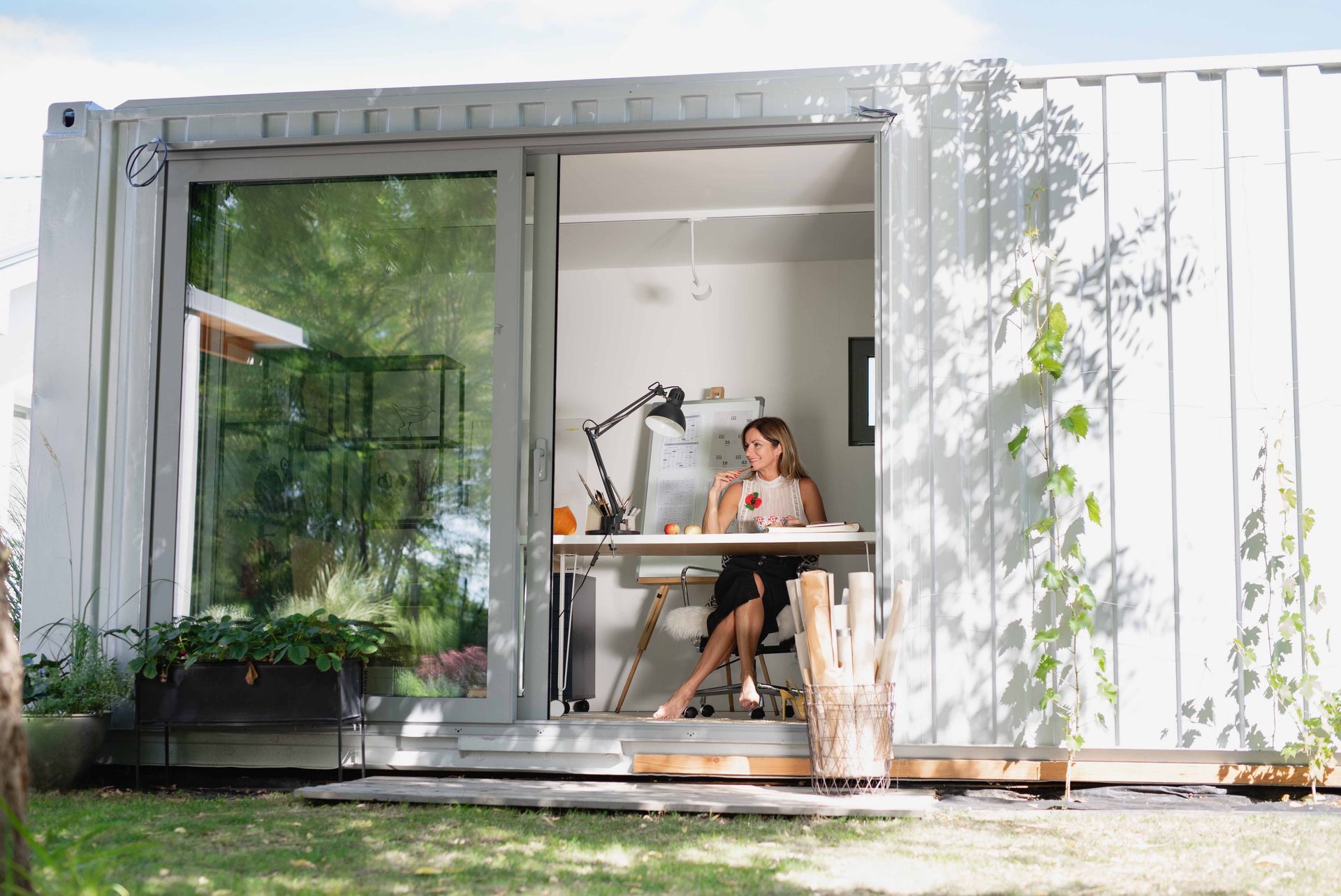 A person sits at a desk working inside a white shipping container office with a large glass sliding door.