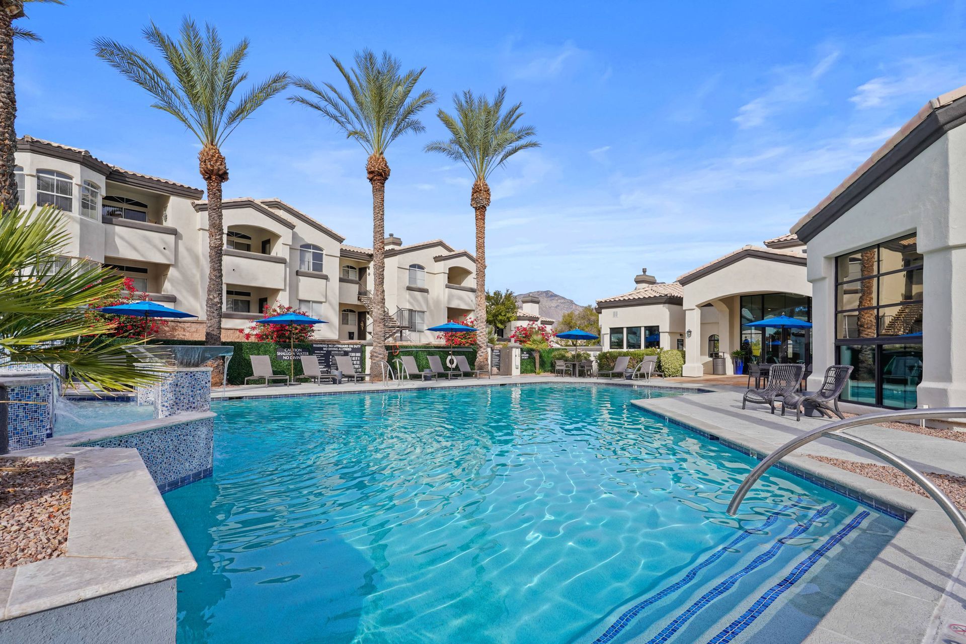 Outdoor community pool with palm trees, lounge chairs, and blue umbrellas.