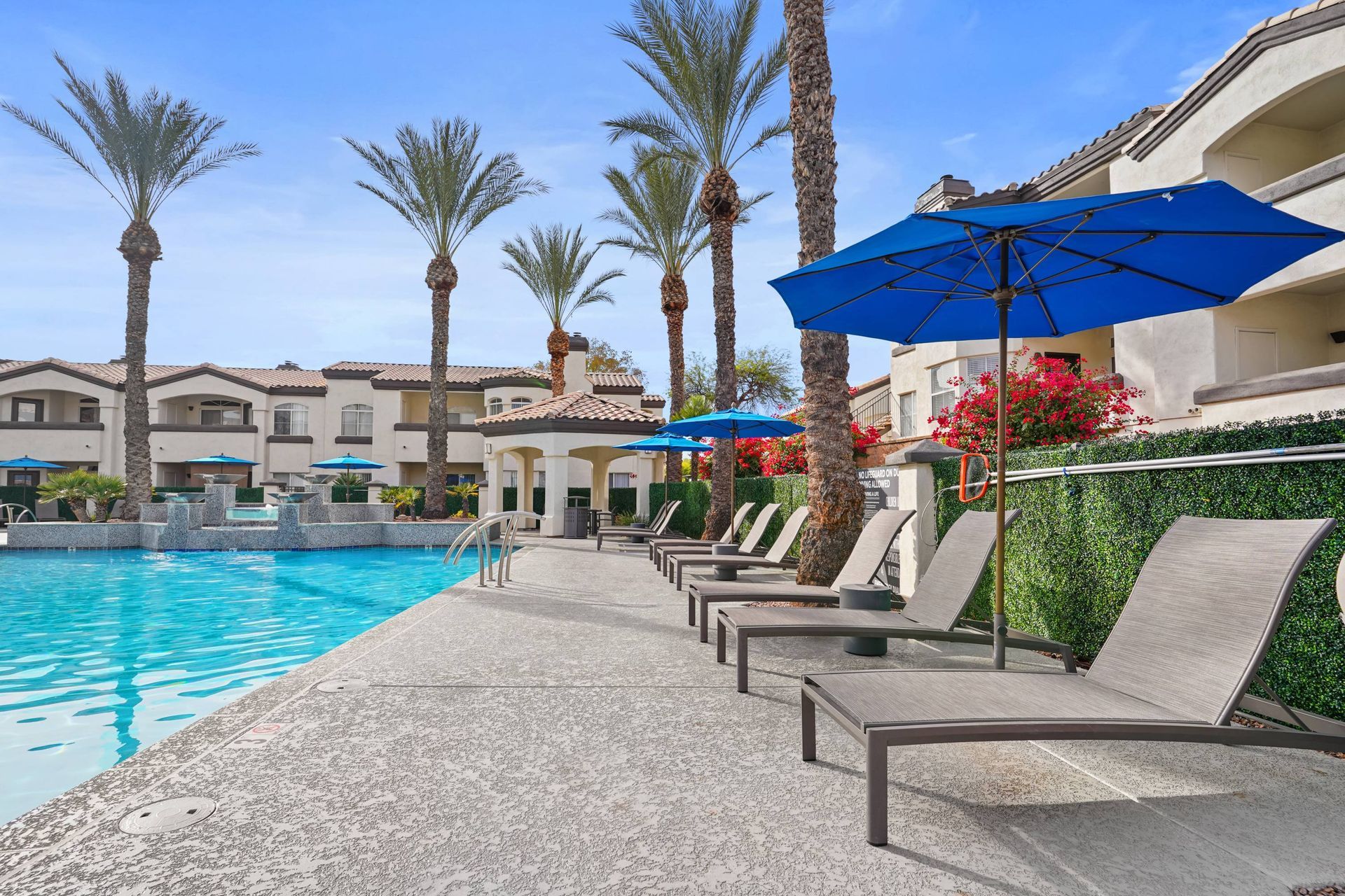 Outdoor pool deck with lounge chairs, blue umbrellas, and palm trees.