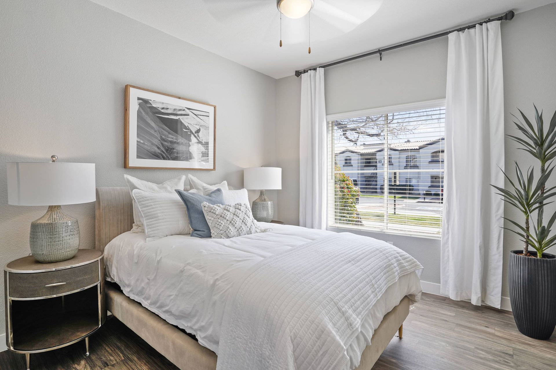 Bedroom in an apartment with a white bed, two nightstands, a lamp, window blinds, and a tall plant.