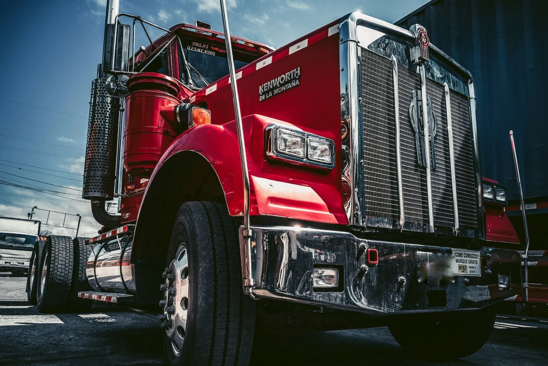 A close-up, low-angle view of a shiny, bright red Kenworth semi-truck parked outdoors under a blue sky.