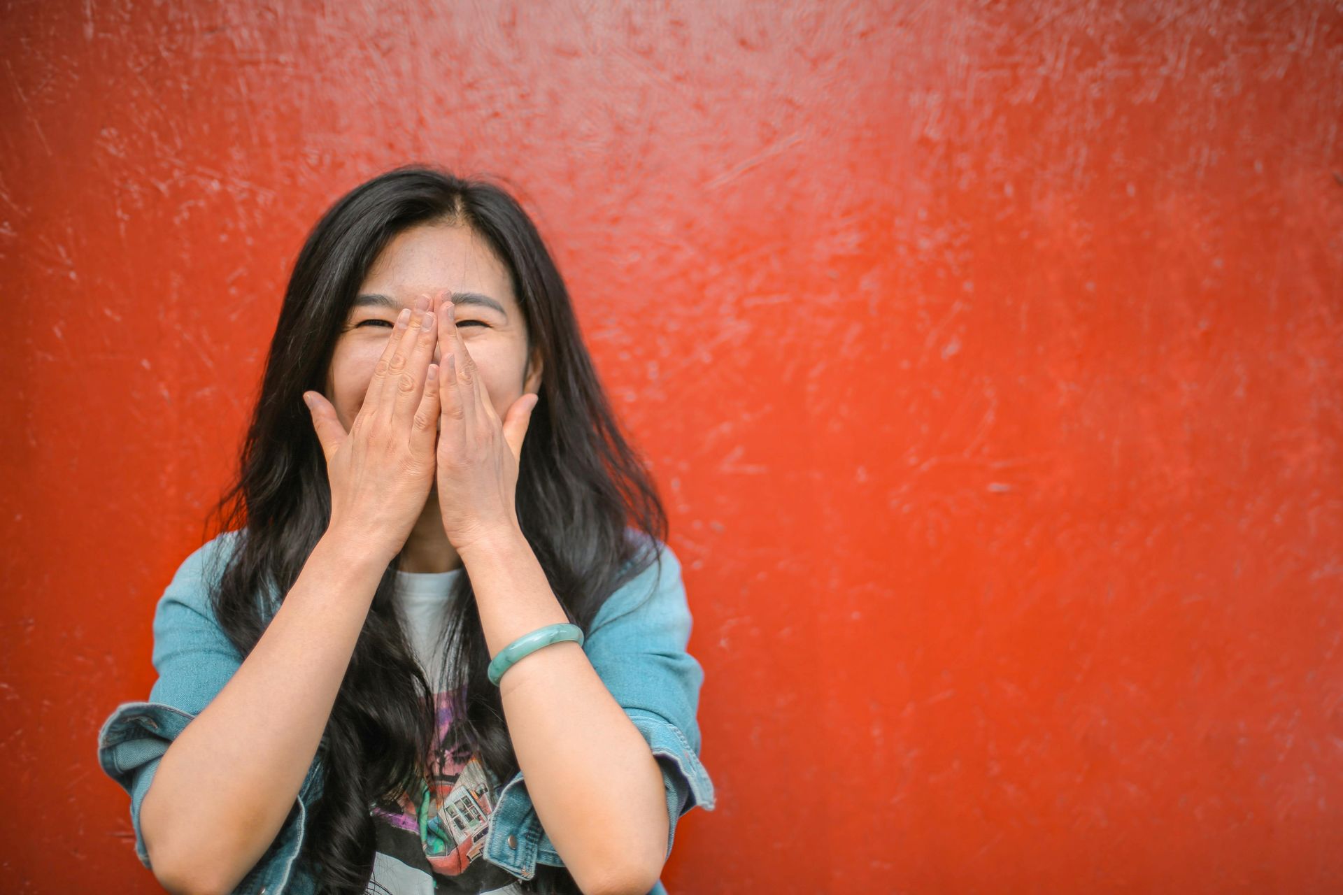 Hong Kong Woman laughing, covering face with hands in front of a red wall. | 扣稅年金