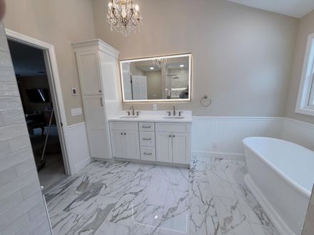 Bright white bathroom with double vanity, chandelier, marble floor, and freestanding tub.