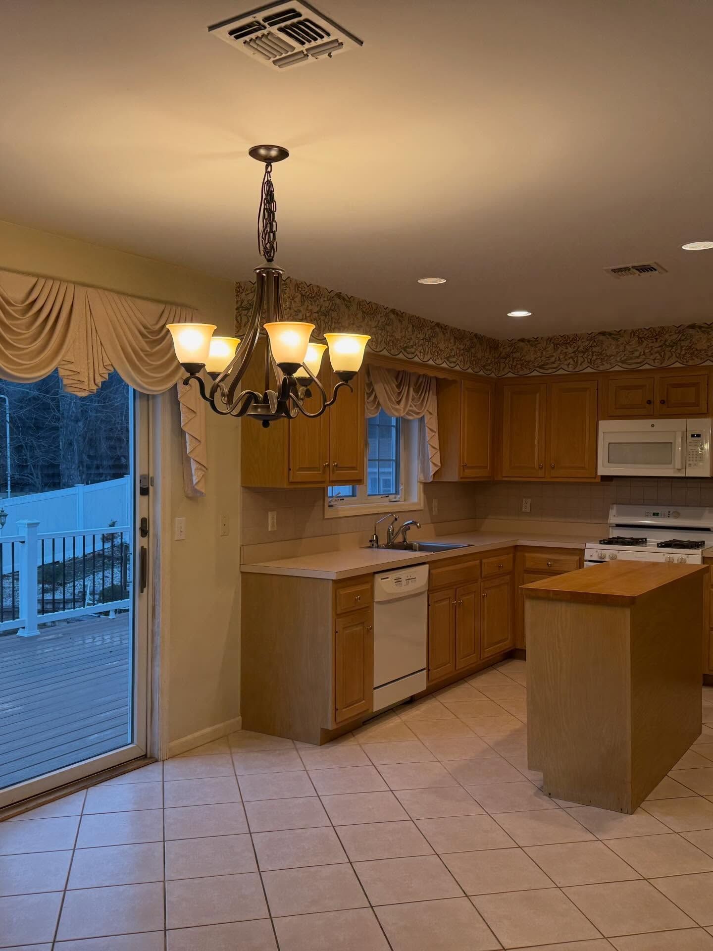 Empty kitchen with wood cabinets, white appliances, tiled floor, chandelier, and sliding glass door.