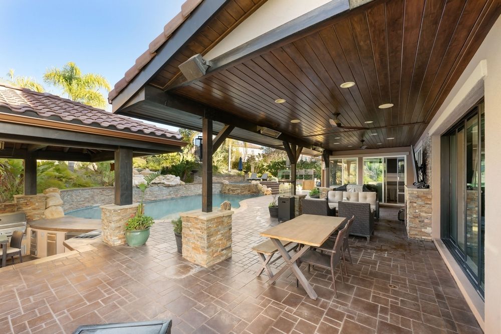 Covered patio with brick flooring, stone columns, and a picnic table beside a house.