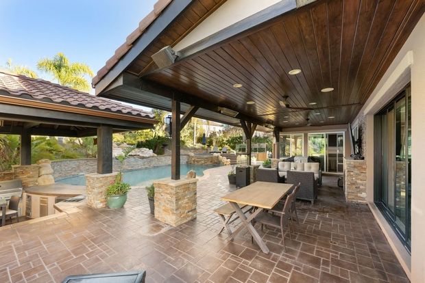 Covered patio with brick floor, wooden ceiling, stone columns, and outdoor dining table by large windows
