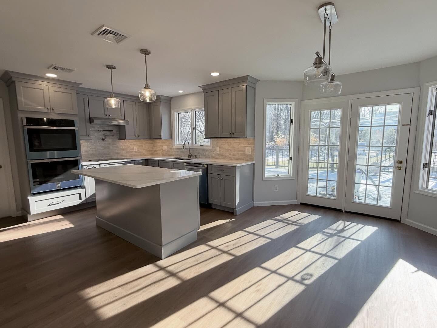 Bright modern kitchen with gray cabinets, white island, and sunlight streaming through glass doors.