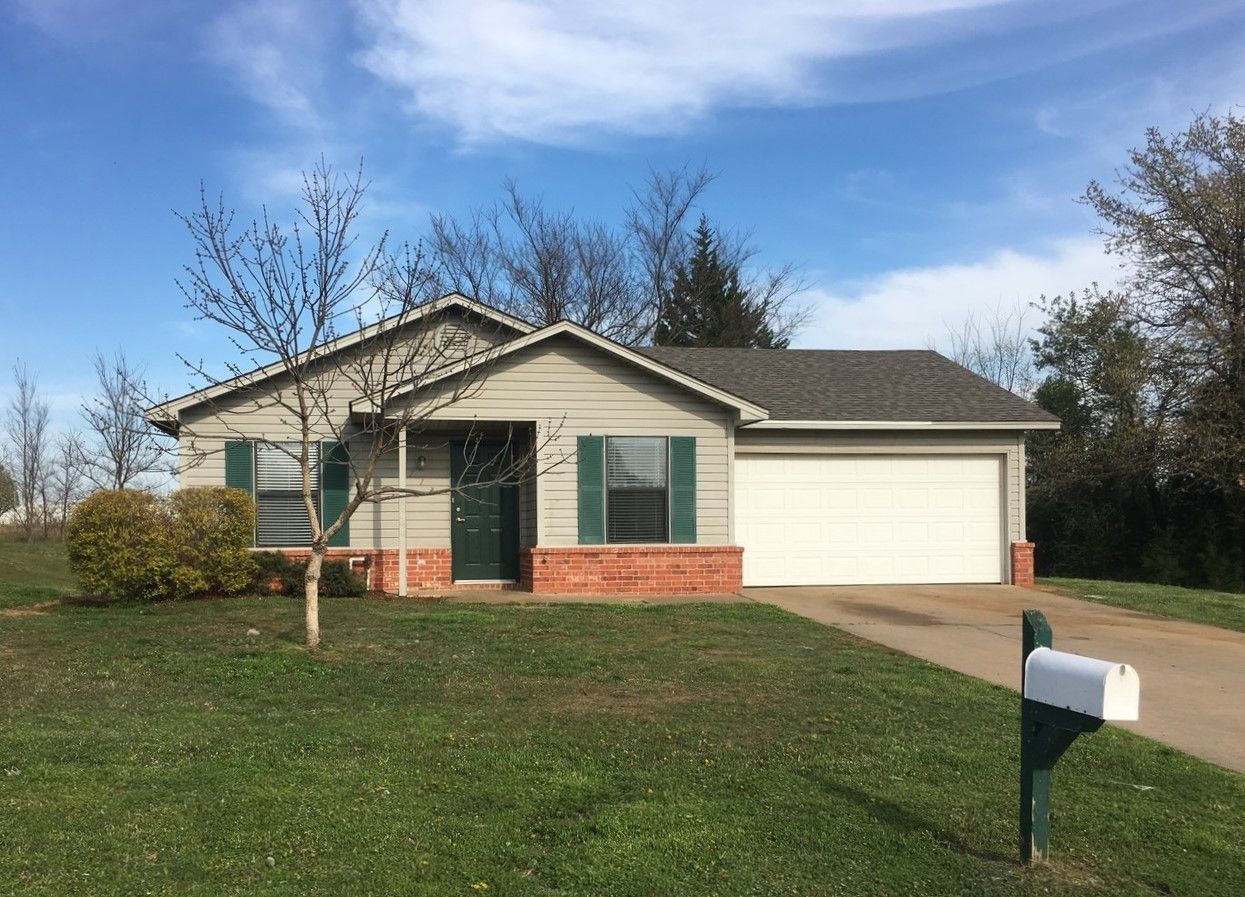 Single-story house with white garage, green shutters, and a mailbox in front yard under a blue sky