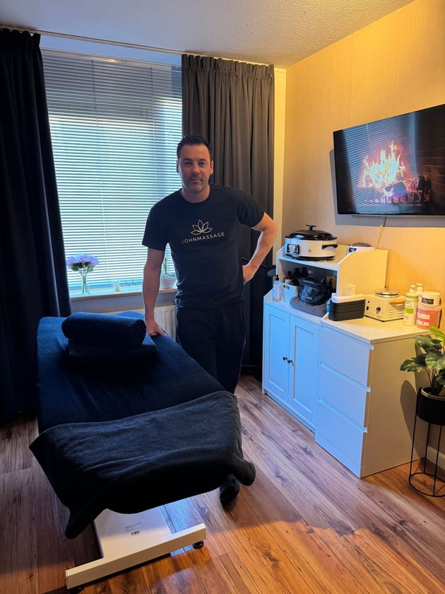 Een man staat in een knusse kamer naast een zwarte stoel, een witte commode en een raam met jaloezie&euml;n.
