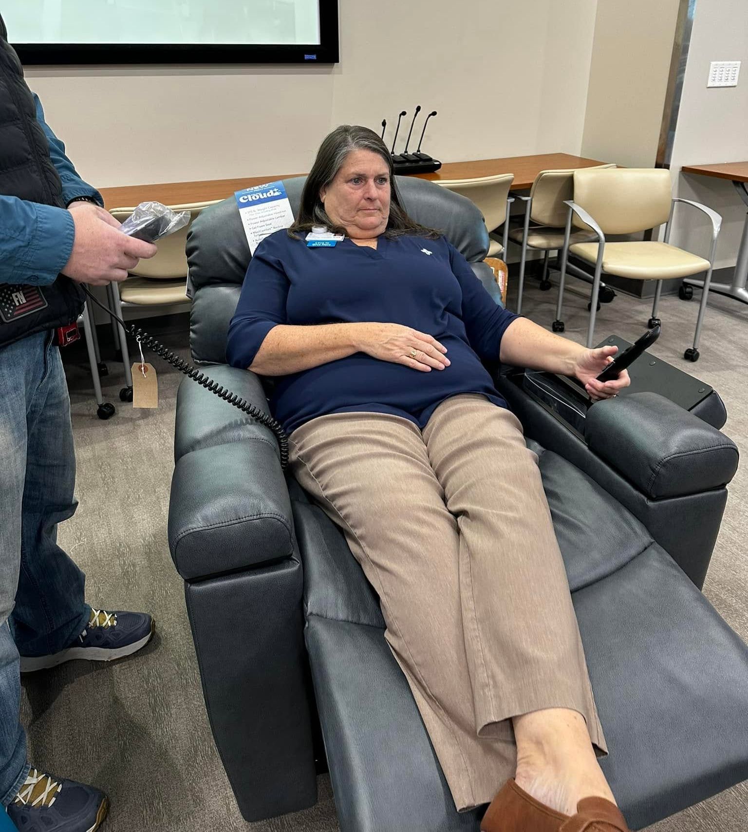 Woman reclines in a dark massage chair in a room with tables and chairs, holding a phone.