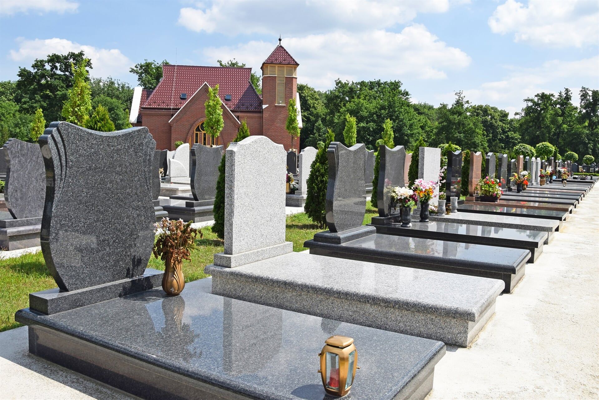 Rows of polished granite gravestones with flowers, framed by a brick chapel and trees.
