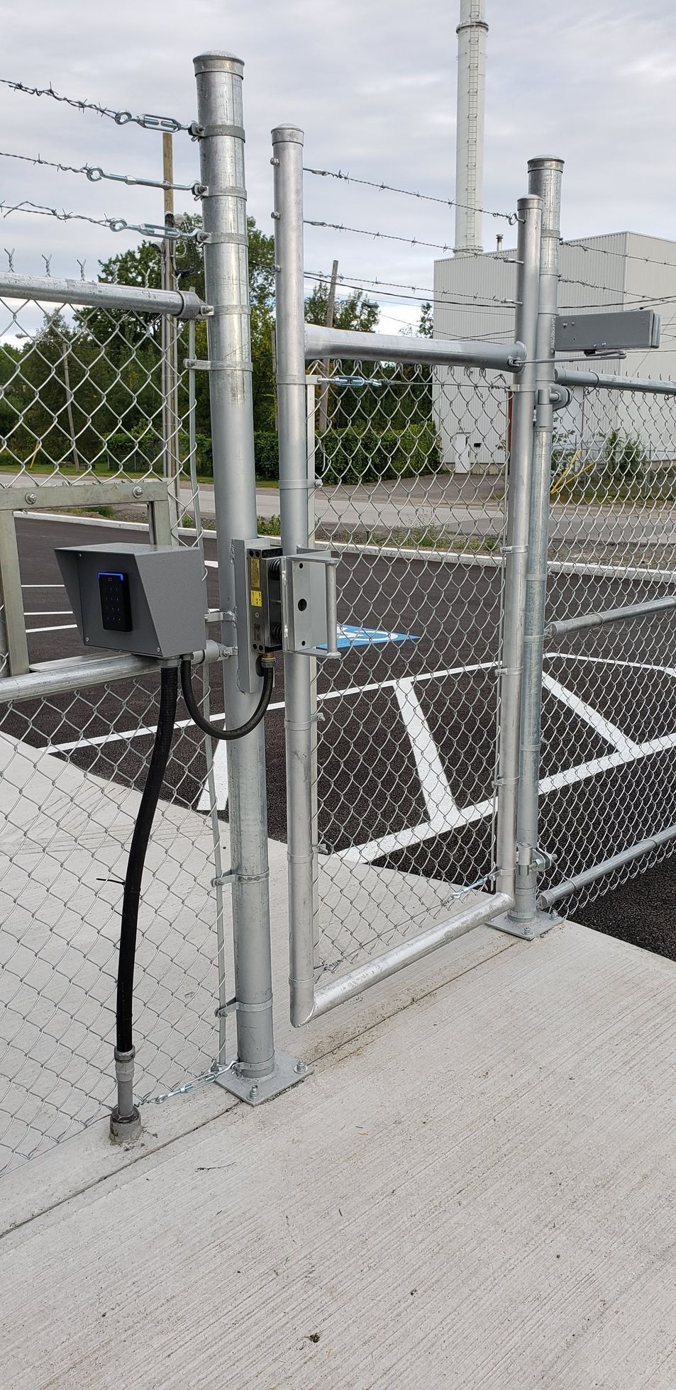 Une clôture à mailles losangées avec un portail et du fil barbelé entourant un parking.