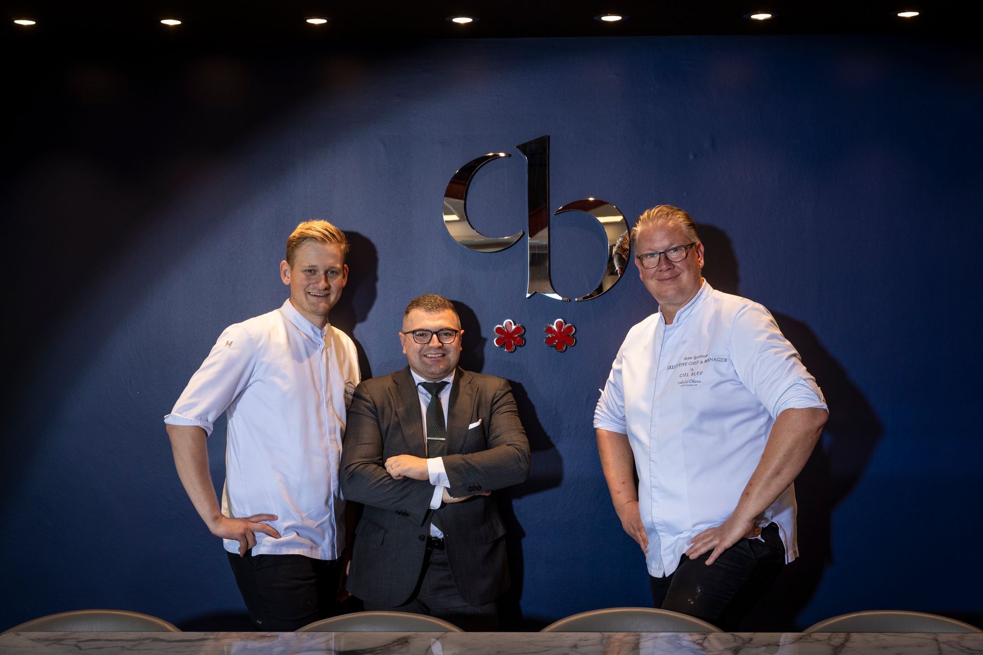 Three people in a modern kitchen. A man in a suit stands between two men in white chef's coats.